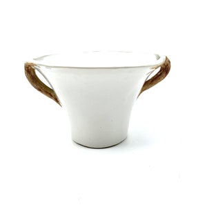 May include: A white ceramic bowl with two brown, textured handles. The bowl has a flared shape and a smooth, glossy finish. The handles are curved and appear to be made of a natural material, adding a rustic touch to the design.