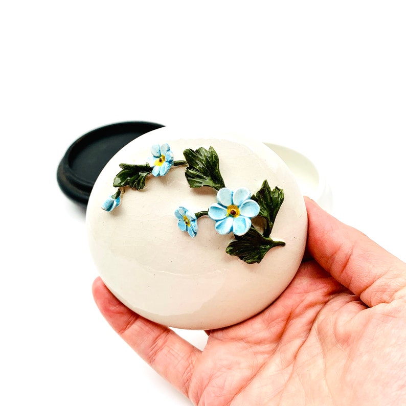 May include: A white ceramic trinket box with a floral design. The box is decorated with blue forget-me-not flowers and green leaves.