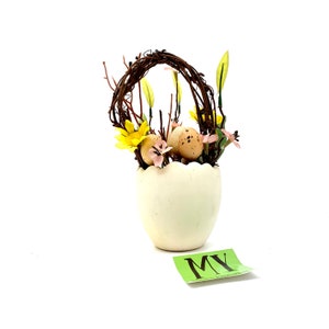 Può includere: Un vaso di ceramica bianco a forma di uovo con un nido di ramoscelli e fiori gialli e rosa. Il vaso è pieno di uova finte.