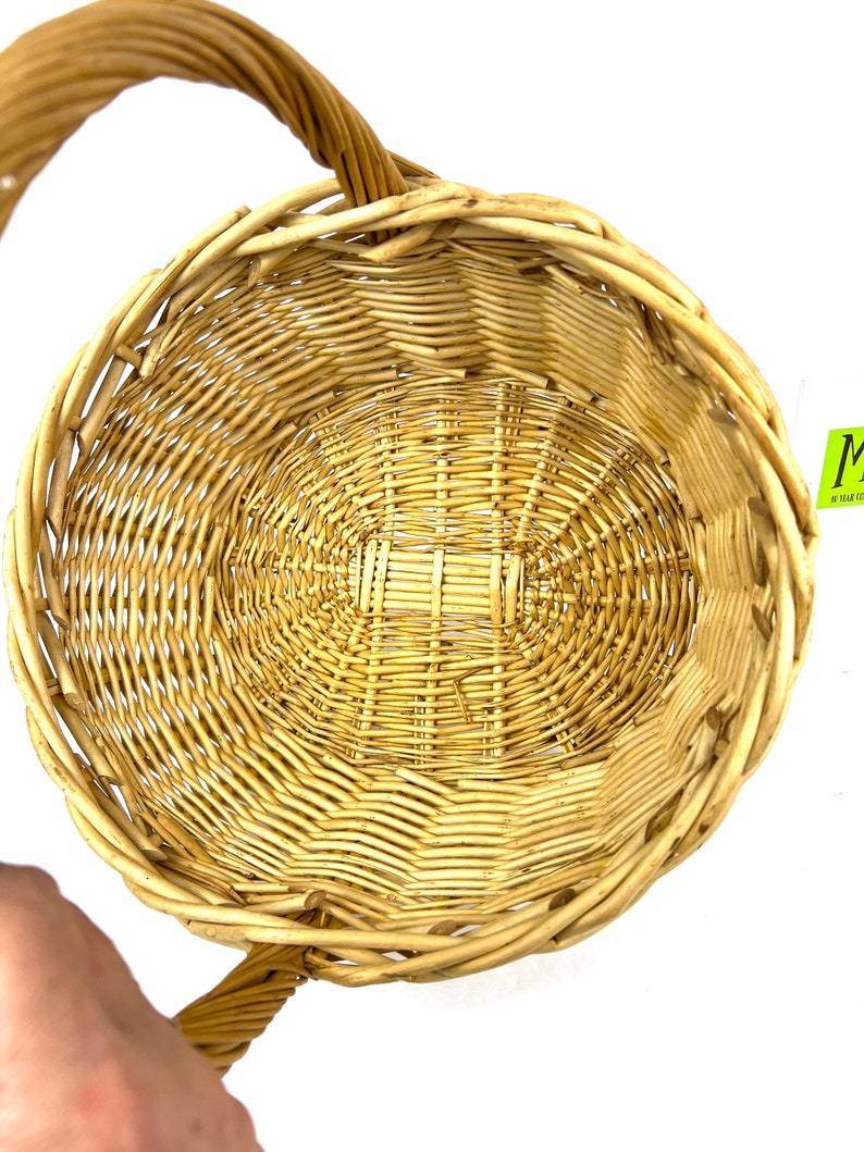 May include: A woven wicker basket with a single handle. The basket is light brown and has a round shape.
