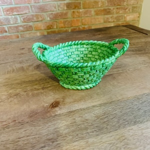 May include: A green woven basket with two handles. The basket is sitting on a wooden surface.
