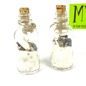 May include: Two small glass bottles with cork stoppers filled with white sand and seashells. The bottles are tied with twine.