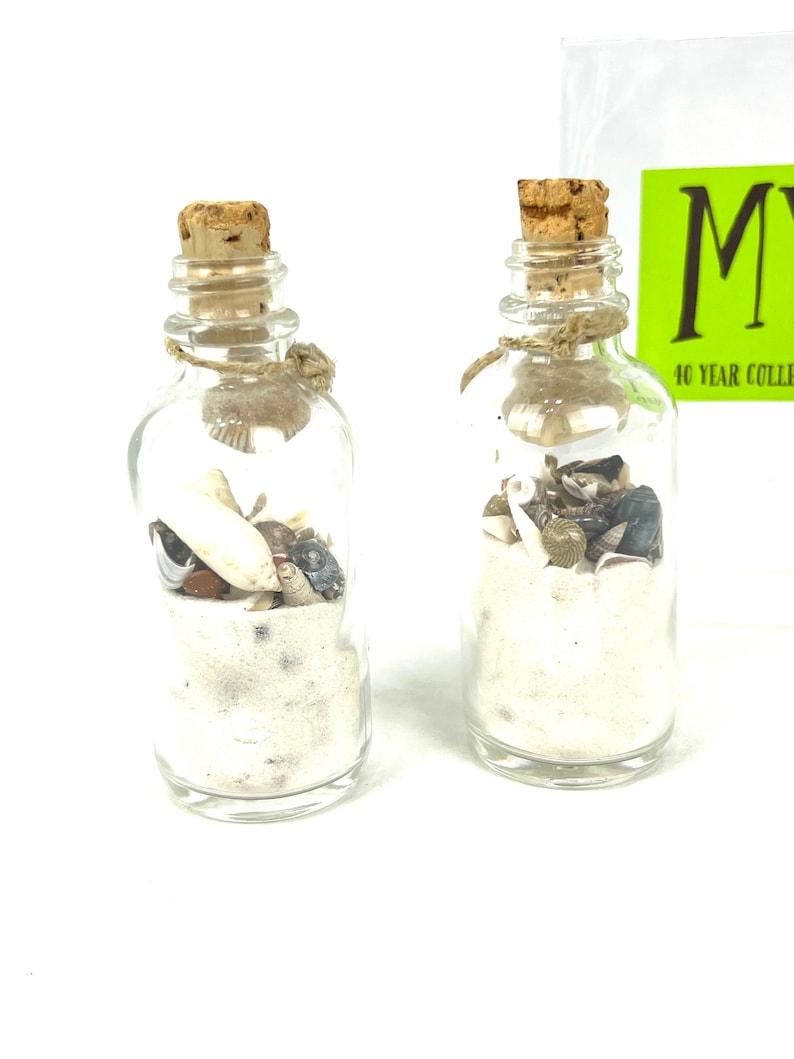 May include: Two small glass bottles with cork stoppers filled with white sand and seashells. The bottles are tied with twine.