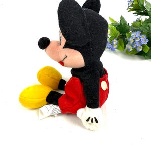 May include: A plush Mickey Mouse toy in a seated position. The toy features a black head and ears, a pink face, red shorts, yellow shoes, and white gloves. The toy is a classic Disney character.