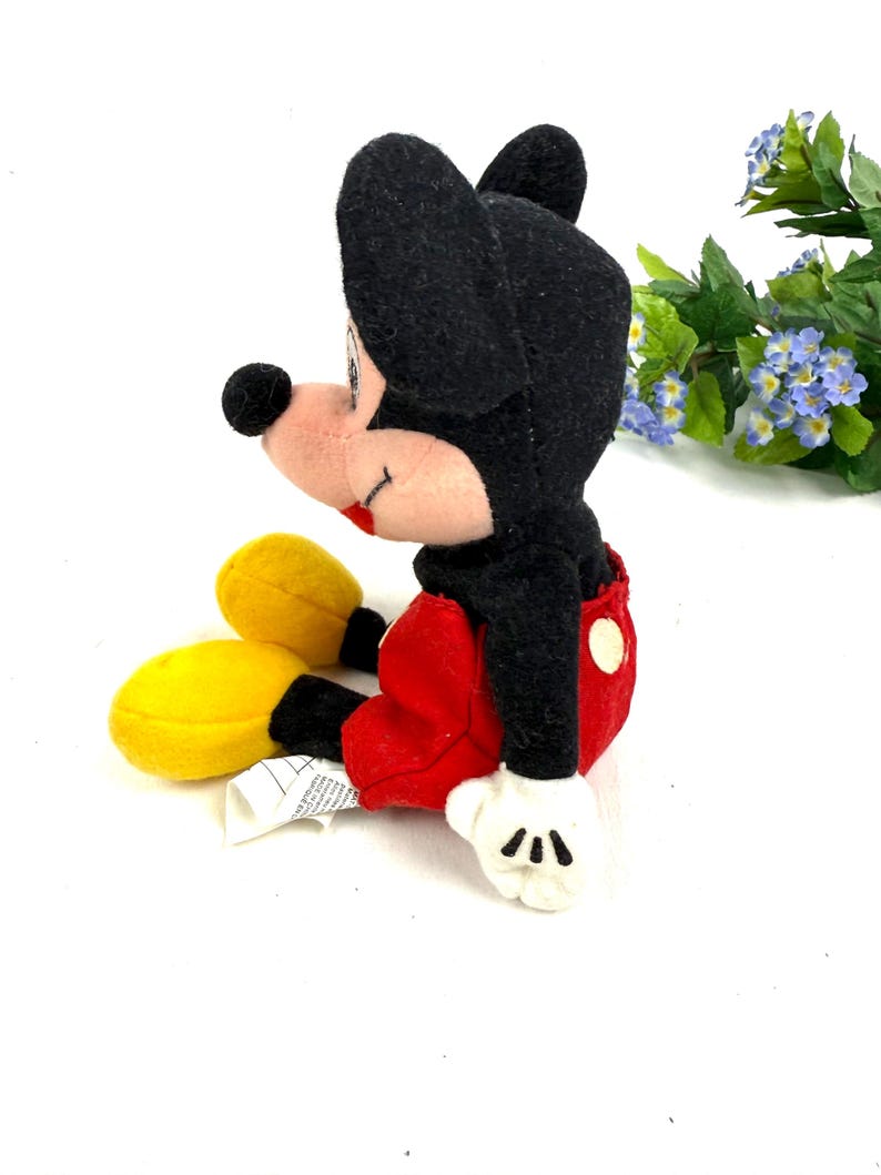 May include: A plush Mickey Mouse toy in a seated position. The toy features a black head and ears, a pink face, red shorts, yellow shoes, and white gloves. The toy is a classic Disney character.