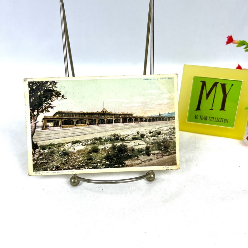 May include: A vintage postcard depicting a sepia-toned image of a long building with arched windows, possibly a railway station. The postcard is held in a silver metal display stand. A small yellow frame with the text "MY 40 YEAR COLLECTION" is also present.