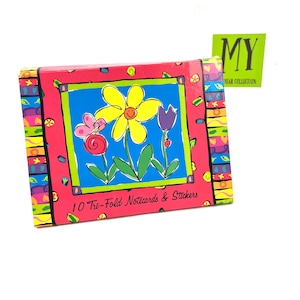 May include: A pink and blue box with a colorful flower design and the text "10 Tri-Fold Notecards & Stickers". The box has a green sticker with the text "MY YEAR COLLECTION".