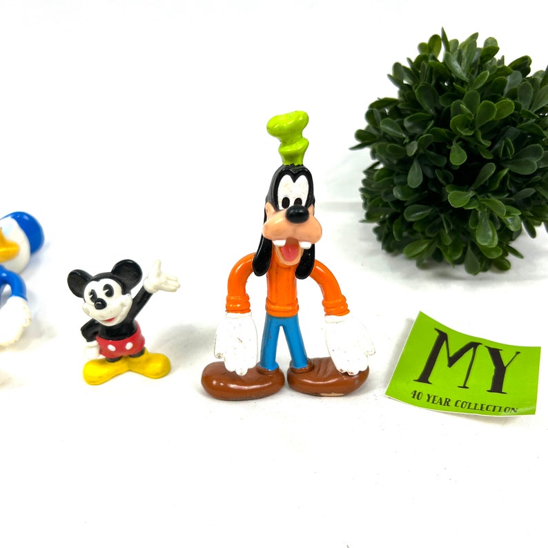 Może przedstawiać: Zestaw trzech wintażowych plastikowych figurek postaci z Disneya. Myszka Miki ma na sobie swoje charakterystyczne czerwone spodenki i ż&oacute;łte buty. Goofy ma na sobie zielony kapelusz, pomarańczową koszulę, niebieskie spodnie i brązowe buty. Kaczor Donald ma na sobie niebieski marynarski str&oacute;j.