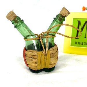 May include: Two small green glass bottles with cork stoppers are tied together with twine and wrapped in a woven straw basket. The bottles are in a V-shape and the basket is red and brown.