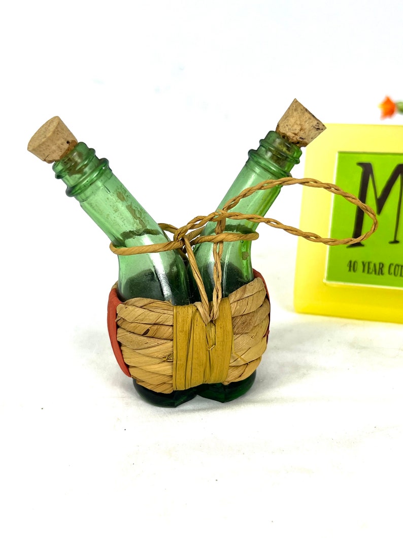 May include: Two small green glass bottles with cork stoppers are tied together with twine and wrapped in a woven straw basket. The bottles are in a V-shape and the basket is red and brown.