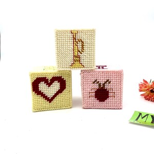 May include: Three small, square, fabric boxes with embroidered designs. The boxes are stacked on top of each other. The top box is white with a gold embroidered letter "I". The middle box is yellow with a red embroidered heart. The bottom box is pink with a red embroidered apple.