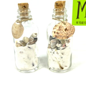 May include: Two small glass bottles filled with white sand and seashells. The bottles have cork stoppers and are tied with twine. The bottles are on a white background.