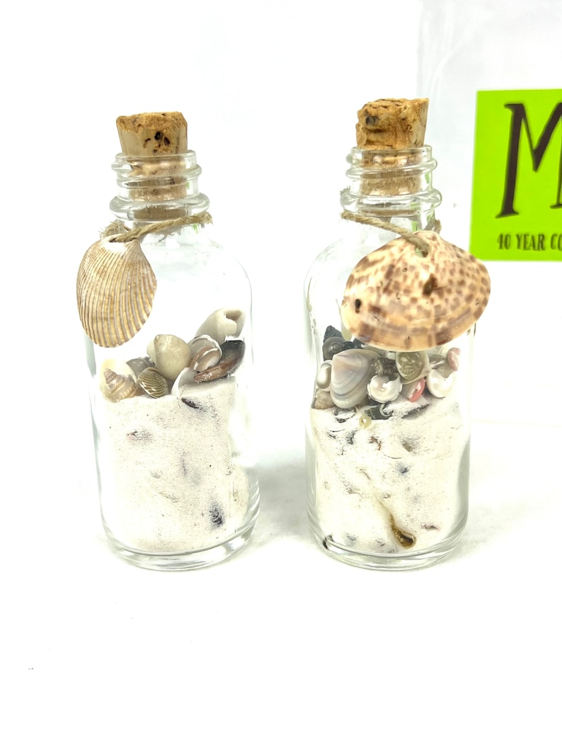 May include: Two small glass bottles filled with white sand and seashells. The bottles have cork stoppers and are tied with twine. The bottles are on a white background.