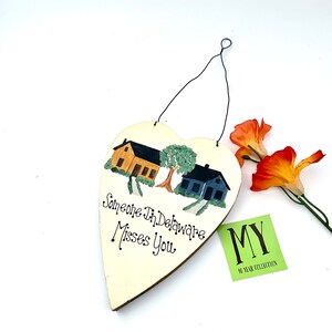 May include: A white heart-shaped wooden sign with a painted scene of two houses and a tree. The sign reads "Someone in Delaware Misses You".
