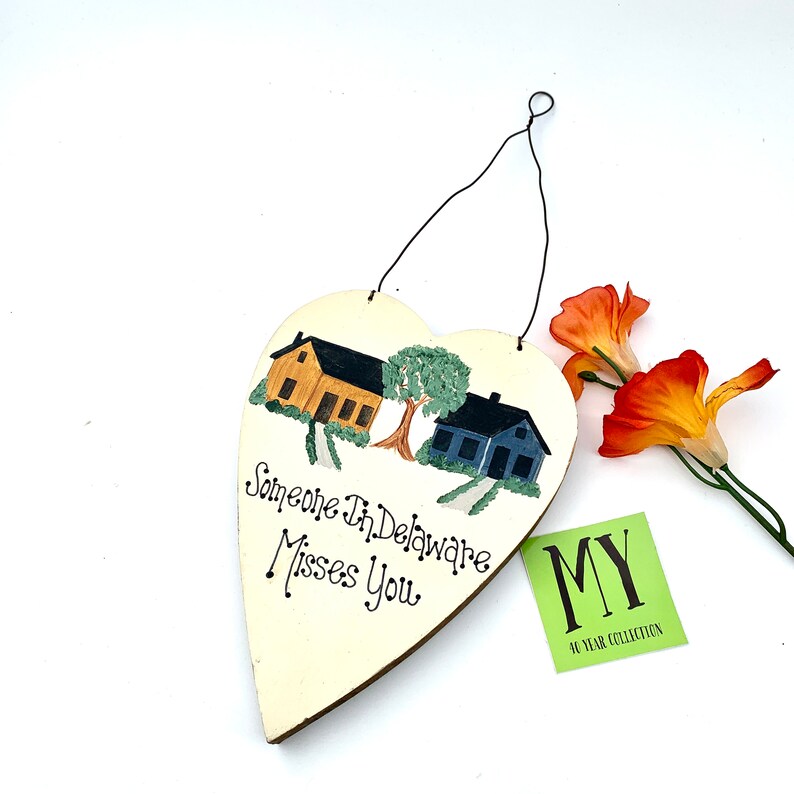 May include: A white heart-shaped wooden sign with a painted scene of two houses and a tree. The sign reads "Someone in Delaware Misses You".