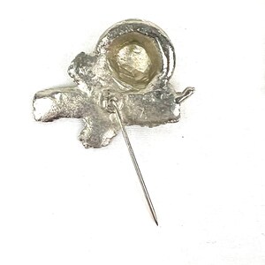 May include: Silver-toned metal pin with a stylized design of a knot or bow. The pin has a pointed back for attaching to fabric.
