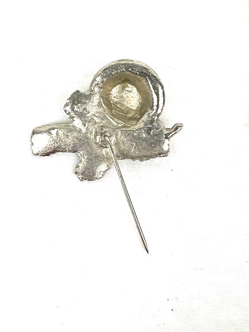 May include: Silver-toned metal pin with a stylized design of a knot or bow. The pin has a pointed back for attaching to fabric.