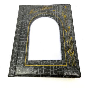 Peut inclure: Un album photo noir avec une texture en faux cuir de crocodile. L'album a une forme rectangulaire avec une grande fen&ecirc;tre en forme d'arche au centre. Des lettres dor&eacute;es et des motifs floraux ornent la couverture. L'album est ferm&eacute; et pos&eacute; &agrave; plat.