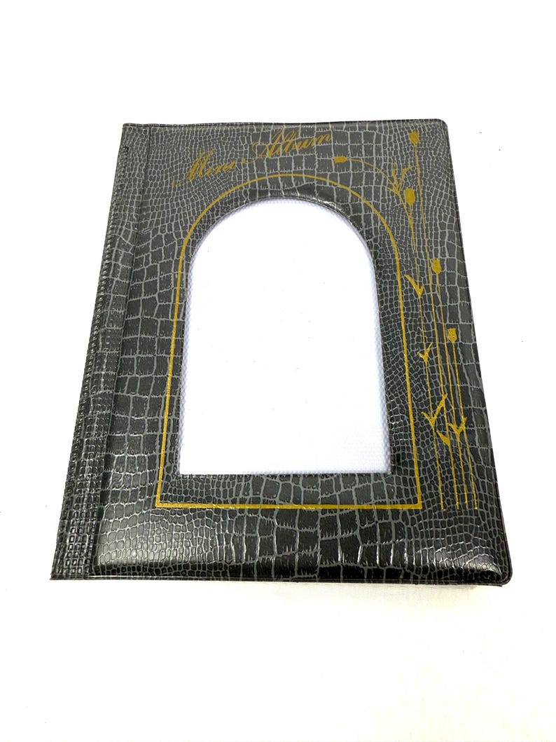 Peut inclure: Un album photo noir avec une texture en faux cuir de crocodile. L'album a une forme rectangulaire avec une grande fen&ecirc;tre en forme d'arche au centre. Des lettres dor&eacute;es et des motifs floraux ornent la couverture. L'album est ferm&eacute; et pos&eacute; &agrave; plat.