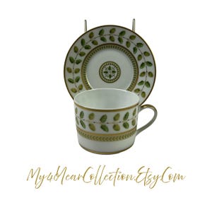 May include: A white porcelain teacup and saucer with a green and gold leaf design. The saucer has a gold rim and the cup has a gold band around the top.