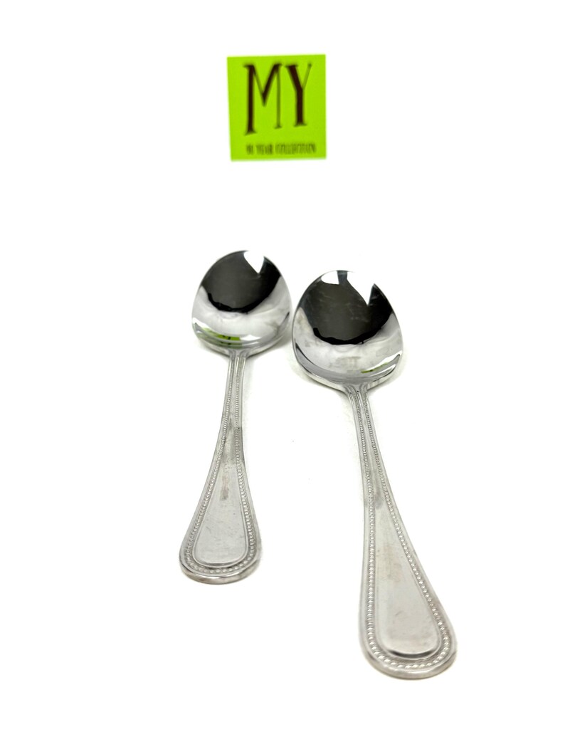 May include: Two silver serving spoons with a slotted design on the spoon head. The handles have a beaded edge and a rectangular design at the end. A green square with the text "MY" is in the background.