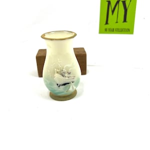 May include: A small, white ceramic vase with a painted scene of a sailboat and seagulls. The vase has a gold rim and a brown wooden base.