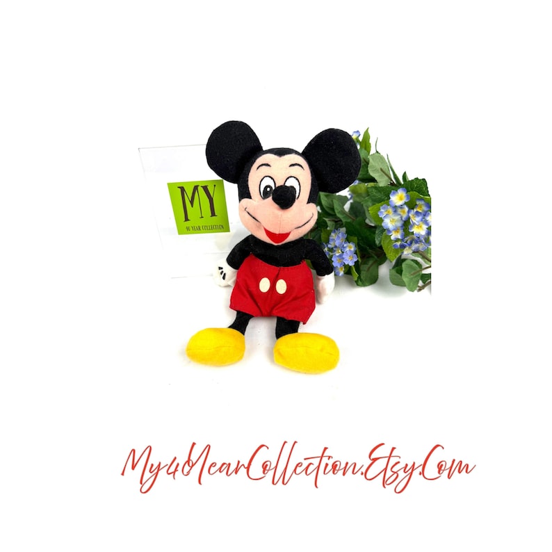 Vintage Mickey Mouse plush toy sitting upright with red shorts, yellow shoes, and flowers in the background.