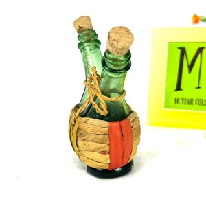 May include: Two small green glass bottles with cork stoppers are tied together with twine and wrapped in a woven straw basket with a red stripe.