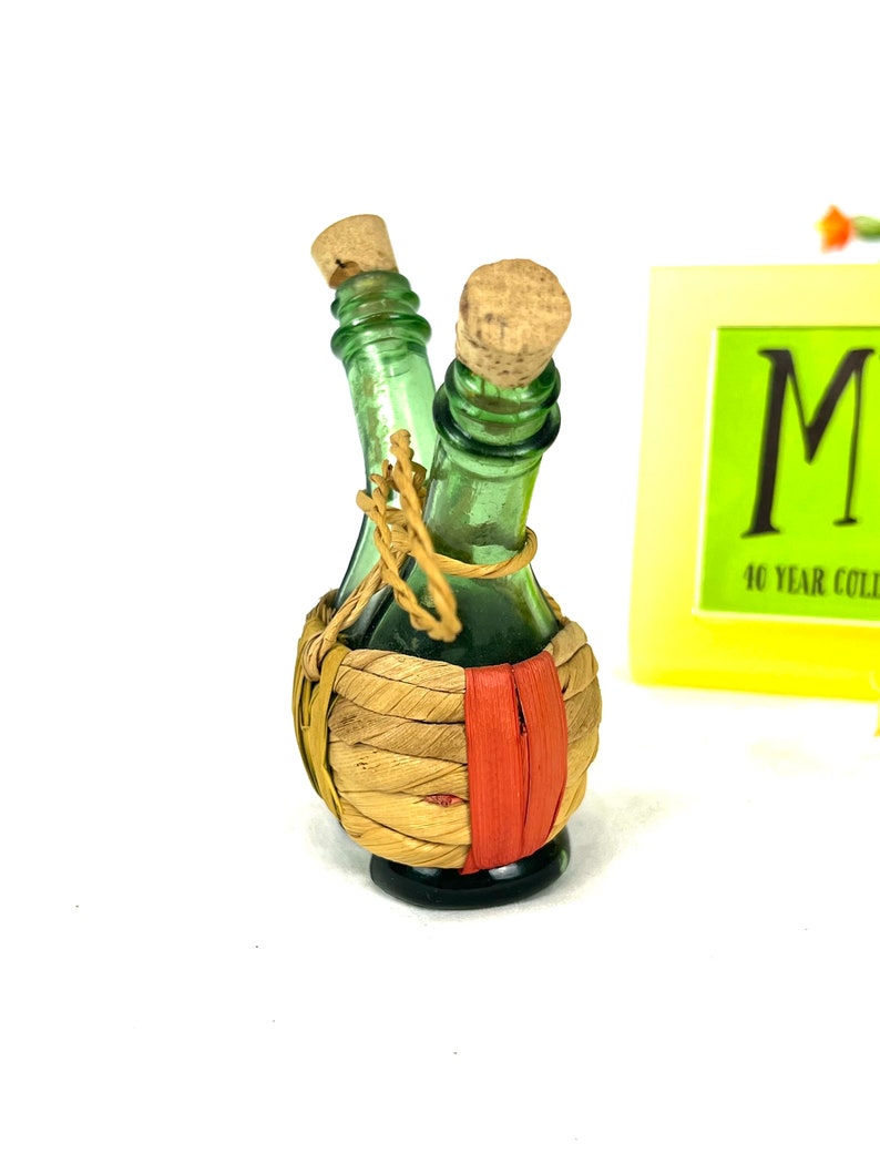 May include: Two small green glass bottles with cork stoppers are tied together with twine and wrapped in a woven straw basket with a red stripe.