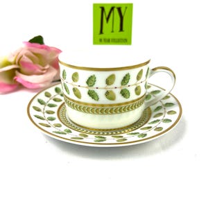 May include: A white porcelain teacup and saucer set with a green and gold leaf design. The saucer has a gold rim and the cup has a gold band around the top. The cup has a handle and the saucer has a scalloped edge.