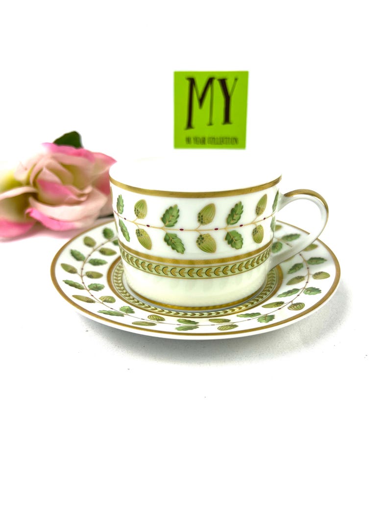 May include: A white porcelain teacup and saucer set with a green and gold leaf design. The saucer has a gold rim and the cup has a gold band around the top. The cup has a handle and the saucer has a scalloped edge.