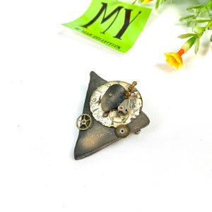 May include: A triangular piece of dark gray clay with a clock face and gears attached. The clock face has a black face with white numbers and hands. The gears are made of brass and are attached to the clock face with small screws.