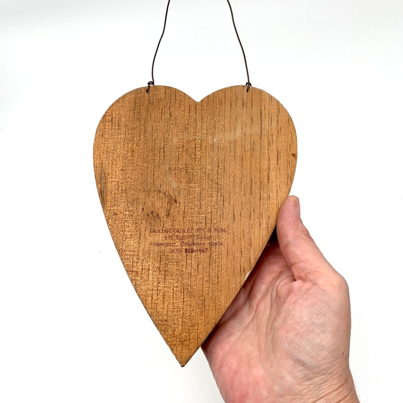 May include: A wooden heart-shaped ornament with a brown finish. The back of the ornament has a label with the text "CELEBRATE LIFE BY GIVING.  +10 TM.  TM.  SERIAL #1004.  Phoenix, Delaware 19804.  (302) 882-1657."