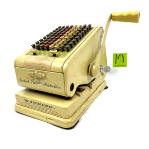 May include: Vintage cream-colored mechanical cash register with a handle and numerous black, red, and gold keys. The register has a drawer labeled "Locked Paymaster Protection" and a warning label. The handle and keys are yellow.