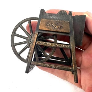 May include: A vintage metal pencil sharpener shaped like a water wheel. The sharpener is painted in a bronze color and has the words "Play Me" on the front. The sharpener is made in Spain.