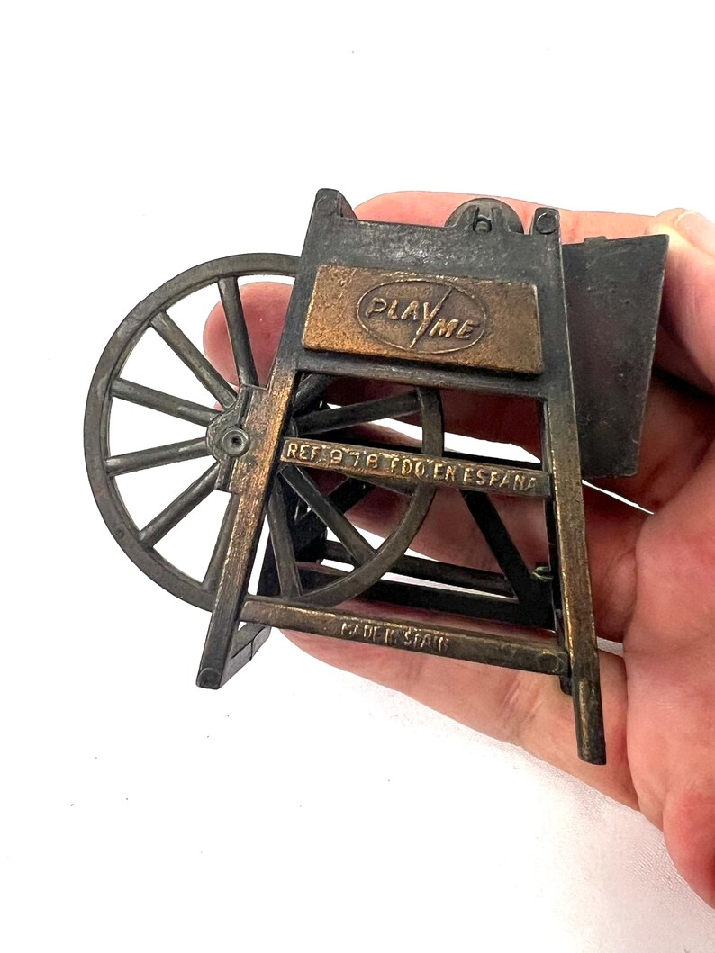 May include: A vintage metal pencil sharpener shaped like a water wheel. The sharpener is painted in a bronze color and has the words "Play Me" on the front. The sharpener is made in Spain.
