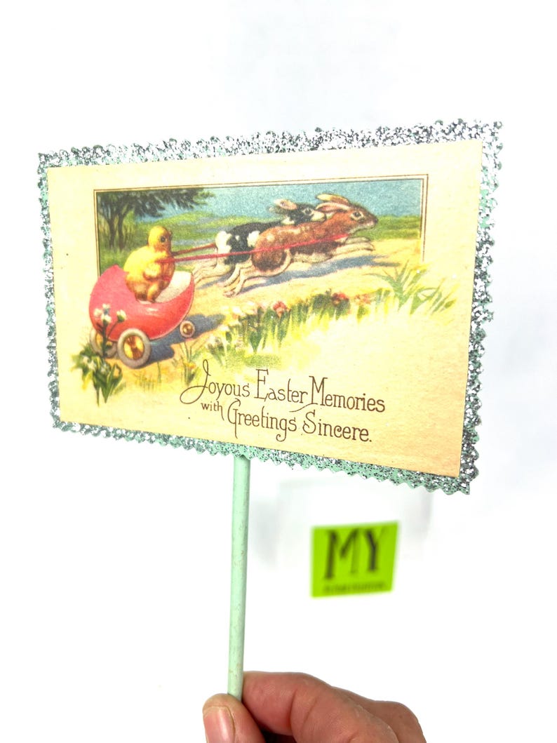 May include: Vintage Easter greeting card on a stick. The card features a chick in a red carriage pulled by two rabbits. The card has a glittery border and the text "Joyous Easter Memories with Greetings Sincere."