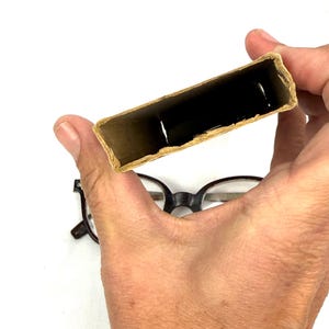 Pode incluir: Uma pessoa segura uma pequena caixa de cart&atilde;o aberta com um interior escuro. A caixa &eacute; retangular e tem uma cor castanha clara natural. Um par de &oacute;culos com arma&ccedil;&atilde;o preta est&aacute; por baixo.