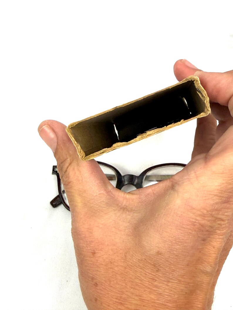 Pode incluir: Uma pessoa segura uma pequena caixa de cart&atilde;o aberta com um interior escuro. A caixa &eacute; retangular e tem uma cor castanha clara natural. Um par de &oacute;culos com arma&ccedil;&atilde;o preta est&aacute; por baixo.