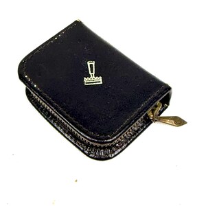 May include: A small, black leather wallet with a gold zipper and a gold emblem on the front. The emblem is a stylized design of a stamp. The wallet is closed and appears to be empty.