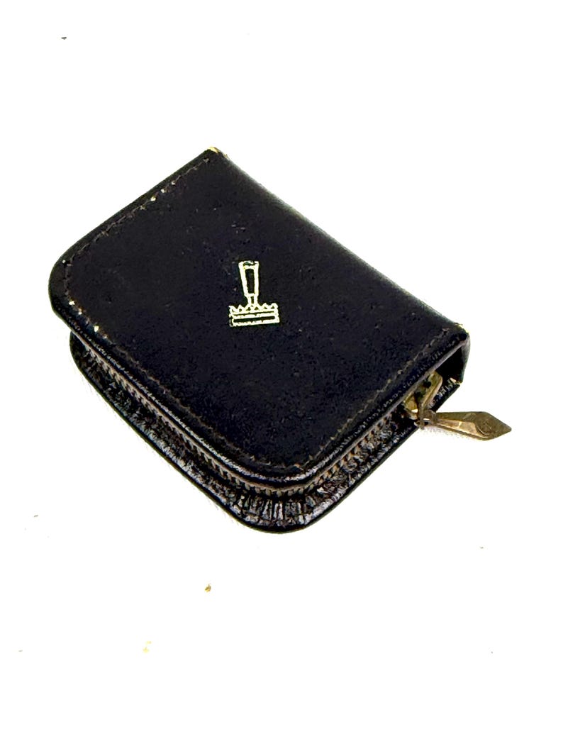 May include: A small, black leather wallet with a gold zipper and a gold emblem on the front. The emblem is a stylized design of a stamp. The wallet is closed and appears to be empty.