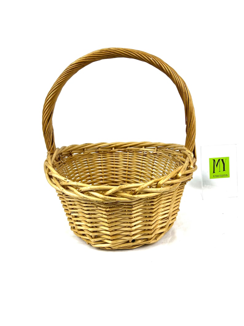 May include: A natural woven wicker basket with a single handle. The basket is light brown in color and has a rounded shape.