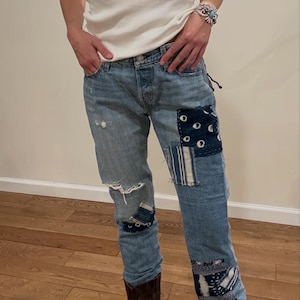 May include: Light blue denim jeans with patches of patterned fabric. The jeans have distressed details and are paired with brown cowboy boots. The patches feature various designs in shades of blue and white.