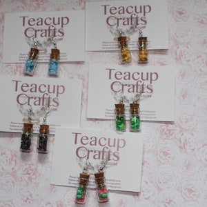 Puede incluir: Cuatro pares de pendientes con pequeñas botellas de vidrio llenas de confeti de colores. Cada par se muestra en una tarjeta blanca con el texto "Teacup Crafts by Louise".