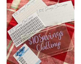 May include: A collection of financial planning tools, including several cards and a clear pouch. The cards feature the text "$10 Savings Challenge" and a grid of dollar signs. A cash breakdown sheet is also included, with a watercolor design.