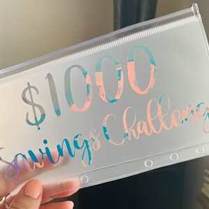 May include: A clear plastic pouch with a zipper closure. The text "$1000 Savings Challenge" is printed on the front in a holographic, iridescent font with shades of blue, pink, and purple. The pouch has three reinforced holes.