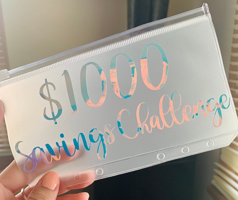May include: A clear plastic pouch with a zipper closure. The text "$1000 Savings Challenge" is printed on the front in a holographic, iridescent font with shades of blue, pink, and purple. The pouch has three reinforced holes.