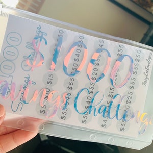 May include: A clear plastic savings challenge envelope with a holographic design. The text "$1000 Savings Challenge" is displayed in a cursive font. The envelope contains a printed sheet with amounts from $5 to $55.