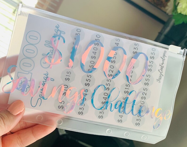 May include: A clear plastic savings challenge envelope with a holographic design. The text "$1000 Savings Challenge" is displayed in a cursive font. The envelope contains a printed sheet with amounts from $5 to $55.