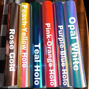 May include: A collection of vinyl rolls in various colors, including rose gold, peach-yellow holo, teal holo, pink-orange holo, purple-blue holo, and opal white. Each roll has a glossy finish and is labeled with its color name in white text.